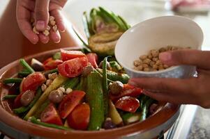 fechar-se do a mãos do uma dona de casa adicionando grão de bico para dentro uma refeição enquanto cozinhando saudável vegetariano refeição dentro tagine foto