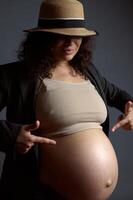 grávida mulher pontos às dela lindo nu grande barriga dentro último trimestre do gravidez, isolado moda cinzento fundo foto