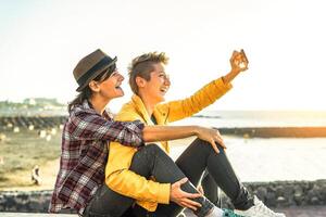 feliz gay casal levando uma selfie com Móvel inteligente telefone Câmera em a de praia às pôr do sol - lésbicas tendo Diversão dentro feriado - homossexualidade, diversidade, férias, viagem, lgbt, tecnologia conceito foto