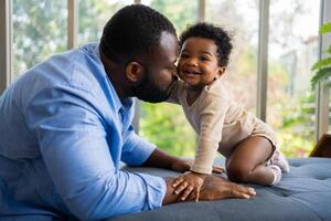 retrato do feliz africano americano Papai com fofa pequeno bebê menina em sofá às casa dentro a vivo sala, carinhoso pai sorridente e divertido dele menina enquanto sentado em a sofá, feliz família foto