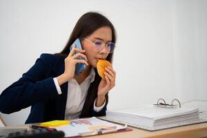 ocupado e cansado empresária comendo pão e leite para almoço às a escrivaninha escritório e trabalhando para entregar financeiro afirmações para uma chefe. sobrecarregado e pouco saudável para pronto refeições, esgotamento conceito. foto