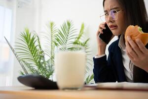 ocupado e cansado empresária comendo pão e leite para almoço às a escrivaninha escritório e trabalhando para entregar financeiro afirmações para uma chefe. sobrecarregado e pouco saudável para pronto refeições, esgotamento conceito. foto