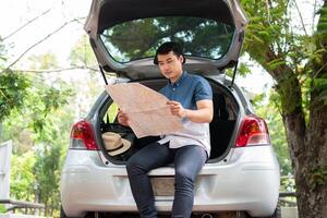 ásia homem sentado costas do carro e segurando mapa para encontrar uma Lugar, colocar para viagem. feliz estrada viagem e período de férias dentro verão dentro a interior. conceito do viagem em lazer e estrada viagem período de férias foto