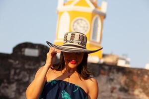 lindo mulher vestindo a tradicional colombiano chapéu chamado sombrero vueltiao às a relógio torre em a histórico ruas do a cartagena de índias murado cidade foto