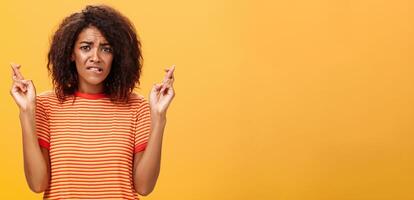 cintura para cima tiro do preocupado perturbado nervoso africano americano mulher com encaracolado Penteado morder mais baixo lábio ansiosamente franzindo a testa cruzando dedos para Boa sorte esperançosamente Rezar para Sonhe venha verdade foto