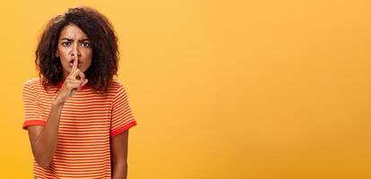 shh Faz não perturbar. retrato do sério mandão e descontente fofa africano americano menina com afro Penteado franzindo a testa calar às Câmera com índice dedo sobre boca proibindo falar alto foto