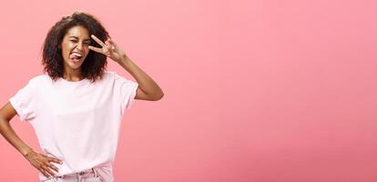 não receoso expressar eu mesmo. alegre carismático africano americano mulher dentro camiseta com afro corte de cabelo mostrando língua de brincadeira e ousadia fazer Paz placa sobre olho e piscando posando sobre Rosa fundo foto