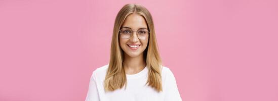 criativo e inteligente boa aparência amigáveis fêmea com bronzeada pele e pequeno espinhas vestindo óculos e branco camiseta sorridente amplamente com perfeito dentes em pé agradável sobre Rosa parede foto