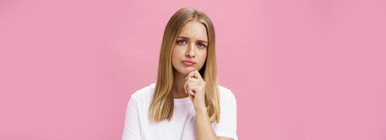 menina voltado para problema pensando ser hesitante enquanto fazer decisão segurando mão em queixo franzindo a testa perseguindo lábios em pé pensativo sobre Rosa fundo dentro branco casual camiseta foto