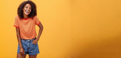retrato do carismático encantador africano americano à moda africano americano fêmea dentro na moda calção e camiseta rindo alegremente desfrutando falando com legal pessoas rindo posando sobre laranja fundo foto