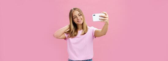 fêmea beleza blogueiro levando selfie postar Novo Veja on-line. retrato do encantador bronzeada jovem mulher dentro camiseta tocante cabelo suavemente puxar mão com Smartphone perto face levando foto sobre Rosa parede