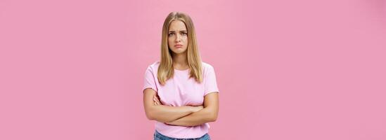 triste inveja e desapontado sombrio fofa jovem mulher com justo cabelo dentro casual camiseta cruzando braços contra peito dentro chateado gesto perseguindo lábios e franzindo a testa olhando preocupado e triste sobre Rosa parede foto