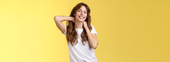 alegre carismático boa aparência cabelo encaracolado mulher alongamento posando amarelo fundo toque pescoço provocante sorridente relaxante perfeito verão período de férias reserva final de semana viagem finalmente descansar debaixo Sol foto