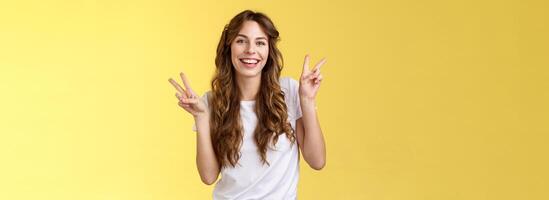 amigáveis pacífico encantador feliz jovem menina encaracolado grandes cabelo mostrar Paz vitória sinais tendo Diversão posando extrovertido animado Veja Câmera divertido rindo sorridente amplamente ficar de pé amarelo fundo foto