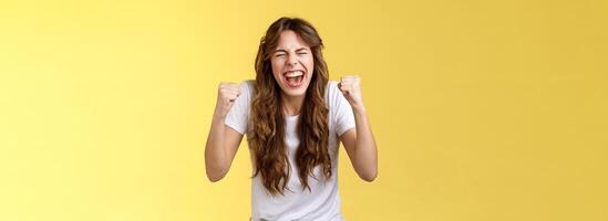 alegre animado feliz entusiasmado menina gritando enraizamento quer ganhar seriamente punho bomba comemorativo satisfeito impressionante sucesso ganhando triunfante alegremente fechar olhos tremendo apertar braços amarelo fundo foto