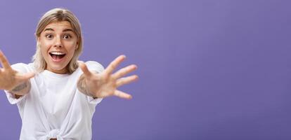 cintura para cima tiro do divertido e animado atraente à moda jovem mulher dentro branco camiseta puxar mãos às Câmera com desejo sorridente emocionados e feliz querendo abraço ou levar alguma coisa sobre roxa fundo foto