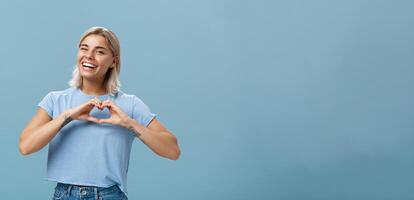 amor relação e emoções conceito. alegre encantador feminino menina com justo cabelo dentro na moda camiseta piscando alegremente sorridente amplamente e fazer coração gesto sobre seio expressando afeição foto