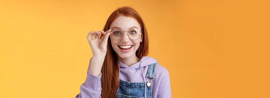 feliz entusiasmado jovem ruiva menina divertido encontrar Fora excelente Lugar, colocar comemoro aniversário em pé alegre animado toque óculos sorridente amplamente branco dentes sorrindo alegria surpreso, laranja fundo foto