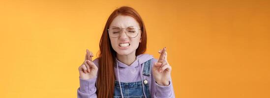 menina desejos ganhar inferior coração suplicante sério apertar dentes franzindo a testa fechar olhos implorando senhor Cruz dedos Boa sorte Rezar fazer sério desejo em pé laranja fundo Sonhe venha verdade foto