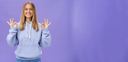 menina obteve tudo debaixo ao controle. Deleitado feliz encantador mulher com justo cabelo e bronzeada pele sem Maquiagem mostrando OK gesto sorridente Assured e satisfeito posando dentro moletom com capuz sobre roxa parede foto