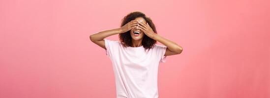 retrato do encantador alegre pele escura brincalhão mulher com encaracolado cabelo dentro camiseta fechamento olhos e contando dez com amplo feliz sorrir jogando ocultar n procurar ou esperando para surpresa sobre Rosa fundo foto