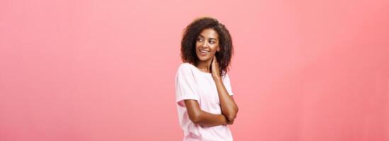 amor dentro ar venha e dizer oi. provocante sensual boa aparência africano americano jovem fêmea aluna com afro Penteado tocante pescoço suavemente girando certo, olhando com curiosidade e desejo sobre Rosa parede foto