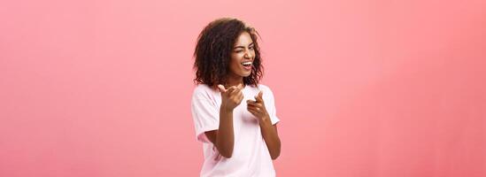 Ei você incrível. brincalhão encantador e feliz despreocupado pele escura na moda menina com encaracolado Penteado piscando e sorridente amplamente fazer dedo arma de fogo mover para Câmera verificação Fora legal equipamento foto