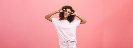 interior tiro do carismático brincalhão pele escura mulher com afro Penteado inclinação cabeça mostrando Paz ou vitória sinais sobre olhos e sorridente sentindo-me divertido arrepiante em impressionante festa sobre Rosa parede foto