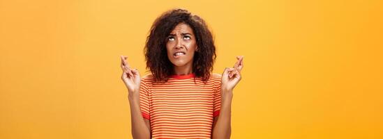 cintura para cima tiro do esperançoso preocupado e ansioso encantador pele escura mulher com afro Penteado dentro na moda listrado camiseta morder lábio nervosamente olhando acima cruzando dedos para Boa sorte enquanto Rezar foto