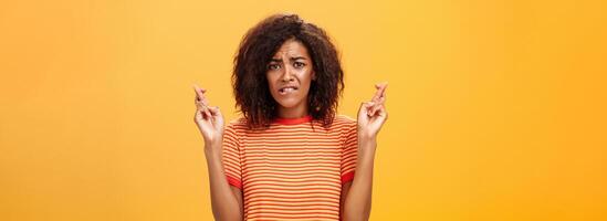 cintura para cima tiro do preocupado perturbado nervoso africano americano mulher com encaracolado Penteado morder mais baixo lábio ansiosamente franzindo a testa cruzando dedos para Boa sorte esperançosamente Rezar para Sonhe venha verdade foto