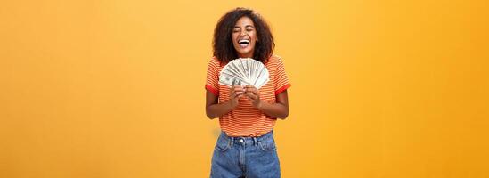 finalmente menina rico. retrato do feliz carismático à moda jovem fêmea modelo recebendo primeiro Forma de pagamento segurando grande quantidade do dinheiro e rindo a partir de alegria ganhando loteria pronto desperdício dinheiro sobre laranja fundo foto