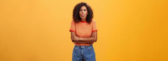 imaturo menina mostrando mau lado do personagem. retrato do infantil ofendido ou descontente jovem afro-americano mulher com encaracolado cabelo mostrando língua cruzando braços em peito sobre laranja fundo foto