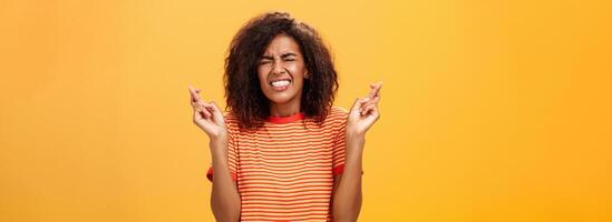 cintura para cima tiro do preocupado preocupado mulher ansioso levar importante trabalho cruzando dedos para Boa sorte aperto dentes e fechamento olhos Rezar sonhando ou fazer desejo sobre laranja fundo foto