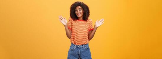 não idéia Faz não Cuidado. descuidado e indiferente calma feliz africano americano mulher com encaracolado cabelo dentro verão roupas levantando Palmeiras e encolhendo os ombros dentro não envolvido pose sorridente despreocupado ser inconsciente foto