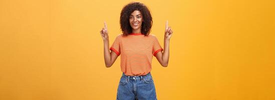 olhando só acima e avançar. otimista ambicioso à moda pele escura fêmea aluna dentro listrado legal camiseta e calção levantando mãos apontando para cima e sorridente amigáveis sobre laranja parede foto