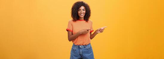 retrato do encantador de aparência amigável afro-americano fêmea colega de trabalho dentro casual equipamento apontando esquerda e sorridente amplamente mostrando perfeito cópia de espaço para clientes contra laranja fundo foto