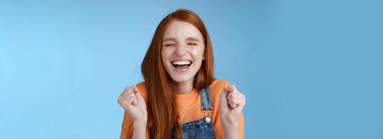 sincero feliz regozijando-se gengibre menina fechar olhos sorridente amplamente dizer sim acenando cerrado punhos alegremente comemoro entrando universidade Sonhe venha verdade ganhando prêmio triunfante alegremente azul fundo foto