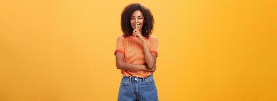 manter segredo dentro Eu Confiar em você. feminino e sensual boa aparência à moda pele escura mulher com encaracolado cabelo dentro na moda equipamento calar às Câmera com Deleitado misterioso Veja se escondendo alguma coisa foto