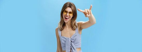 cintura para cima tiro do confiante e ousadia encantador jovem mulher dentro Coincidindo na moda roupas gritando sim mostrando rock-n-roll gesto e olhando às Câmera vestindo legal oculos de sol desfrutando festa ou festival foto