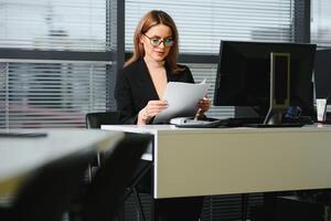 bonito, legal, bonitinho, perfeito mulher sentado às dela escrivaninha em couro cadeira dentro trabalhos estação, vestindo copos, roupa formal, tendo computador portátil e caderno em a mesa foto