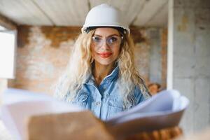 atraente fêmea construção trabalhador dentro capacete. confiante jovem especialista dentro xadrez azul camisa dentro jeans em pé dentro esvaziar sala. interior Projeto e renovação serviço. foto