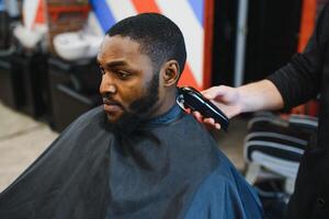 retrato do jovem Preto homem ser aparado com profissional elétrico tosquiadeira máquina dentro barbearia.masculino beleza tratamento conceito. jovem africano cara obtendo Novo corte de cabelo dentro barbeiro salão foto