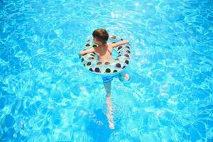 crianças verão período de férias. criança natação dentro piscina. crianças tendo Diversão às Parque aquático. engraçado Garoto em inflável borracha círculo. horário de verão. atrações conceito foto