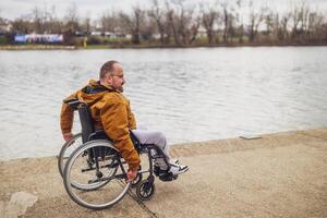 feliz paraplégico deficiente homem dentro cadeira de rodas de a lago. ele é rolando em passarela. foto