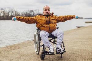 paraplégico deficiente homem dentro cadeira de rodas de a lago. ele é exercício com pesos. foto