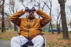 paraplégico deficiente homem dentro cadeira de rodas é usando binóculos ar livre. ele é assistindo pássaros. foto