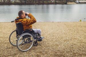 paraplégico deficiente homem dentro cadeira de rodas é usando binóculos ar livre. ele é assistindo natureza de a lago. foto