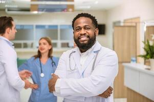 médico sorrisos às Câmera com dele braços cruzado e médico equipe fundo dentro hospital foto