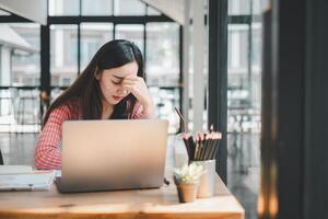 uma jovem mulher parece estressado e cansado enquanto concentrando em dela computador portátil tela dentro uma bem iluminado escritório ambiente. foto