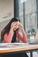 uma jovem ásia mulher parece estressado e cansado enquanto trabalhando em dela computador portátil dentro uma casual escritório contexto. foto